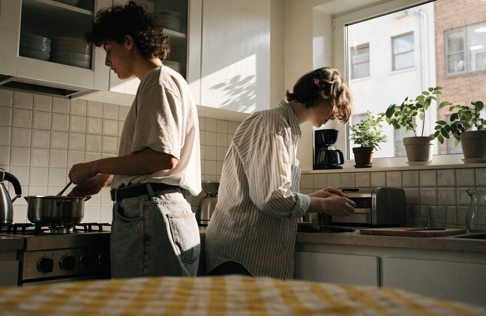 Two residents cooking side by side in the sunlit shared kitchen of a sober living home